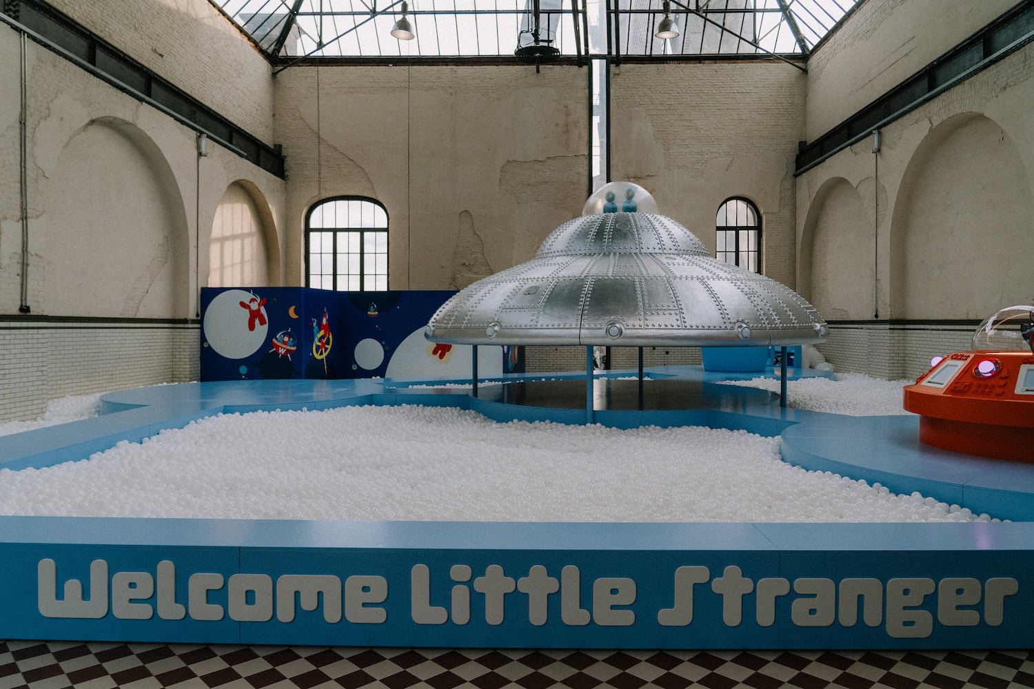 Surreal indoor playground by Walter Van Beirendonck at C-mine, featuring a UFO installation above a ball pit and a “Welcome Little Stranger” sign in a former industrial space.