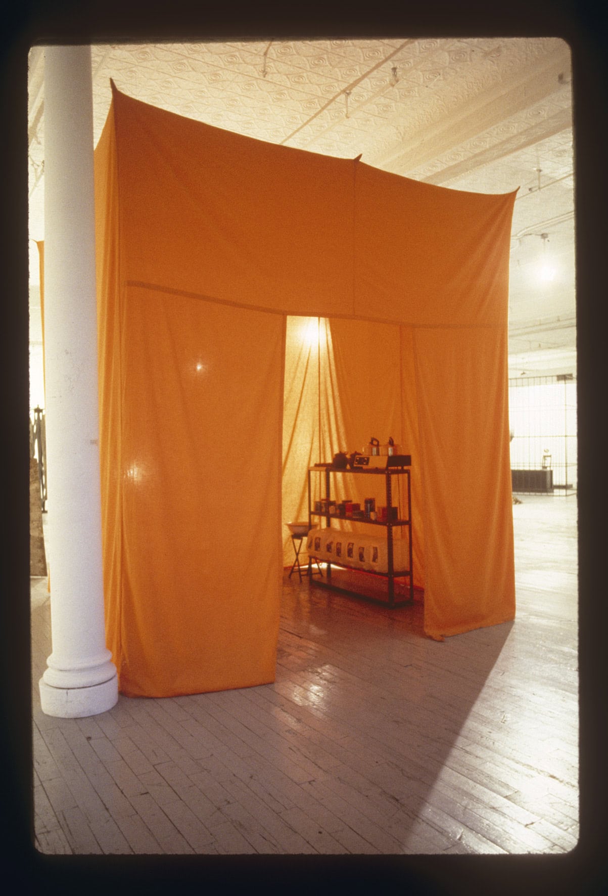 Orange fabric installation forming a tent-like structure inside a gallery, with a partially open entrance revealing shelves with objects and a softly lit interior inviting visitors to enter.