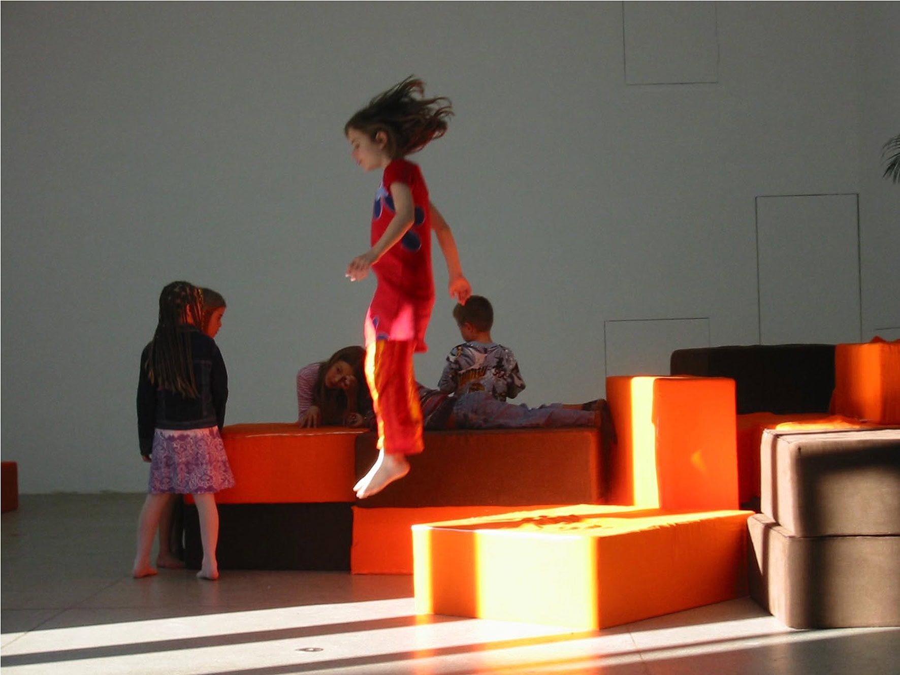 Children interacting with modular orange and brown soft blocks in a contemporary art installation, with one child mid-jump while others sit and explore the space in a minimal gallery setting.