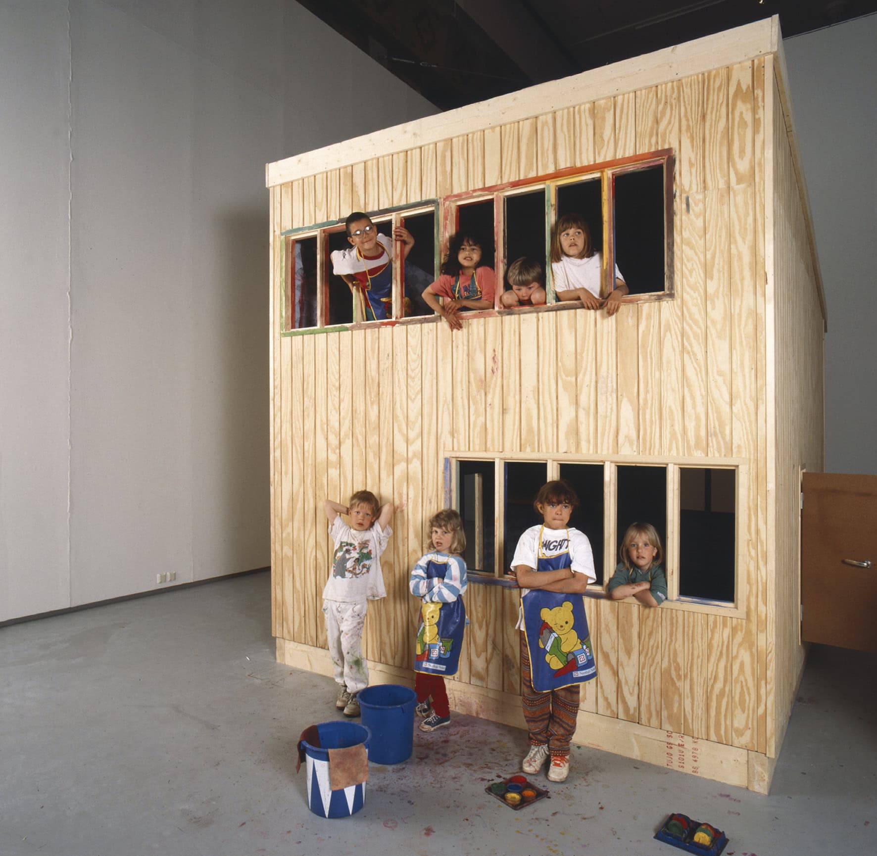 Children gathered around and inside a small wooden house installation in a gallery space, some leaning out of colorful window frames while others stand outside with paint buckets and brushes, suggesting a playful, creative activity.