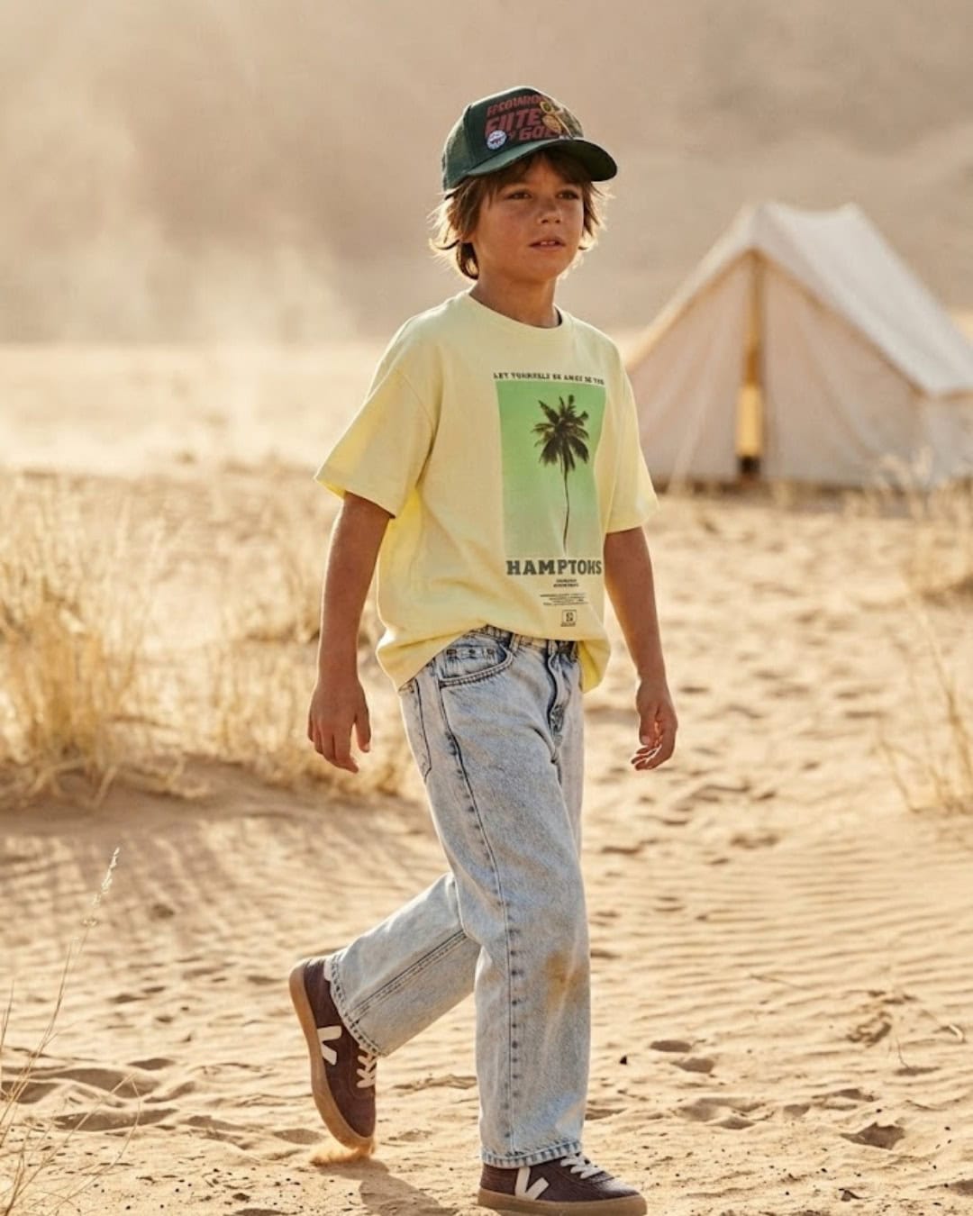 Scimparello Magazine Styling Tip Aries featuring a boy walking through sandy dunes at golden hour, wearing a pale yellow graphic t-shirt, light wash denim jeans, burgundy sneakers and a green cap, desert landscape with tent in the background, warm cinematic light