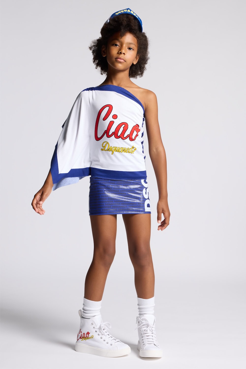 Girl wearing Dsquared2 Kids Ciao Italia capsule collection outfit with one-shoulder logo top, blue mini skirt and white sneakers, SS26 kidswear look.