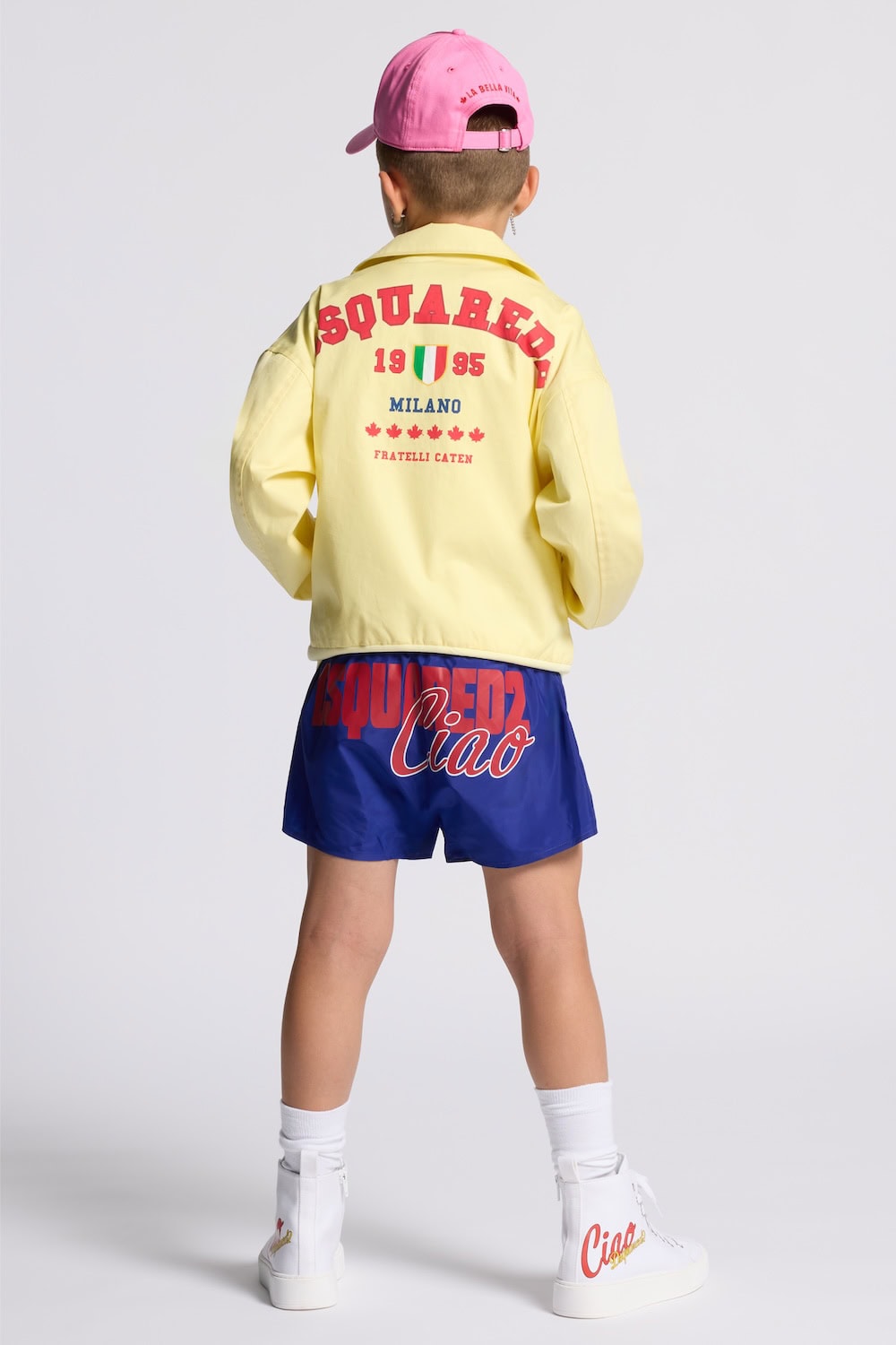 Boy wearing Dsquared2 Kids Ciao Italia capsule collection outfit with yellow jacket featuring Milano logo print, blue shorts with Ciao graphic and white high-top sneakers.