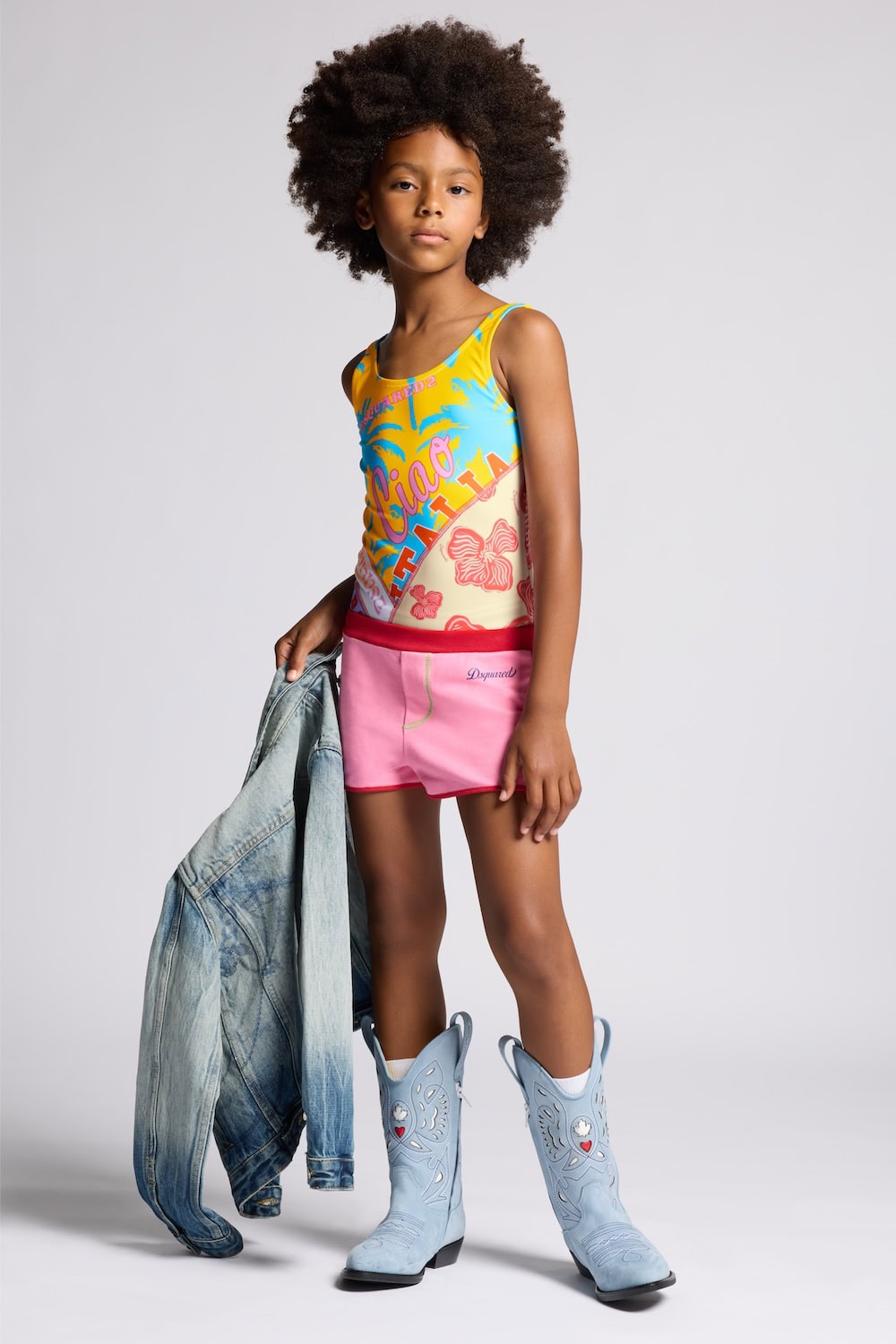 Girl wearing Dsquared2 Kids Ciao Italia SS26 swimsuit with colorful Italian summer print, pink shorts and light blue cowboy boots, holding a denim jacket.