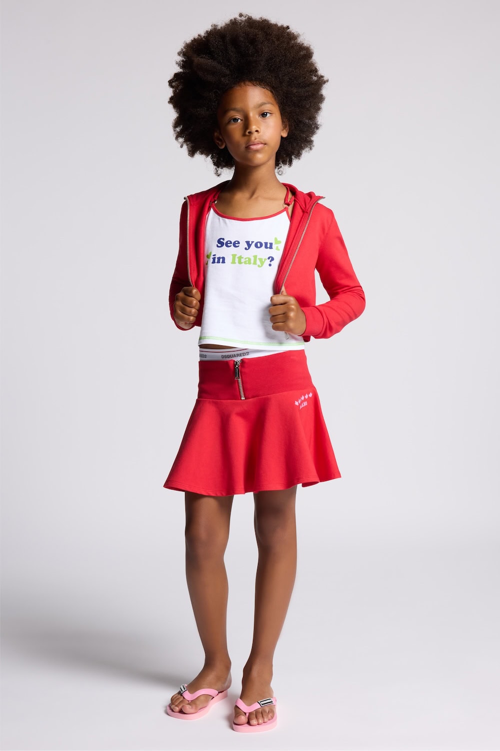Girl wearing Dsquared2 Kids Ciao Italia SS26 outfit with red skirt and hoodie, “See you in Italy?” graphic top and pink flip flops.