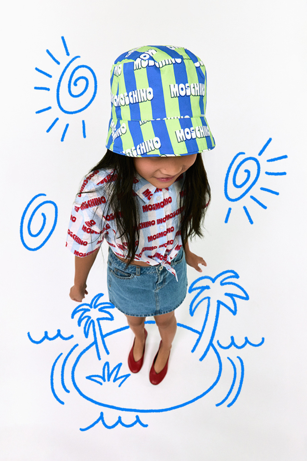 Moschino Baby Kid Teen SS26 look featured in Scimparello Magazine, showing a young girl photographed from above wearing a blue and green striped Moschino bucket hat with logo print, a white short-sleeve shirt covered in red Moschino lettering tied at the waist, a denim mini skirt, and red ballet flats. She stands against a white background decorated with playful blue doodle illustrations of suns, palm trees, waves, and a small island, creating a fun summer holiday mood.