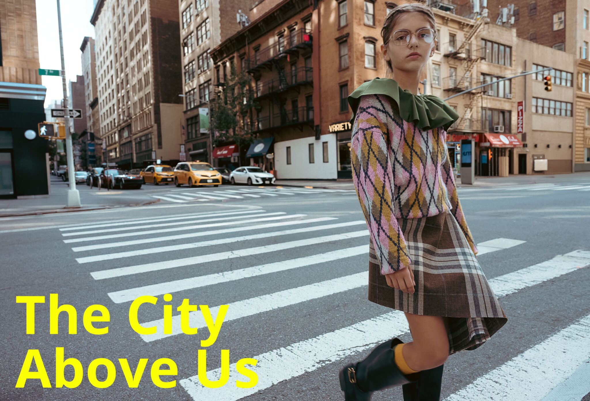Kids fashion editorial The City Above Us for Scimparello Magazine. Girl wearing argyle knit sweater with green ruffled collar and plaid skirt crossing a New York street with yellow taxis and city buildings in the background. Photo by Ilaria and Silvia Cianciaruso.
