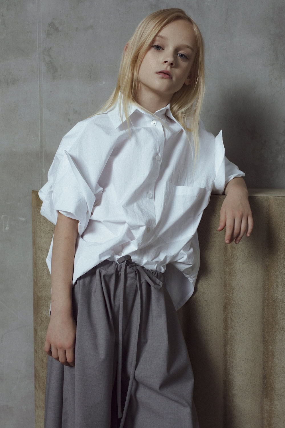 Portrait of girl in crisp white oversized shirt and high-waisted grey trousers with tie belt, UNLABEL SS26 collection, Scimparello Magazine.