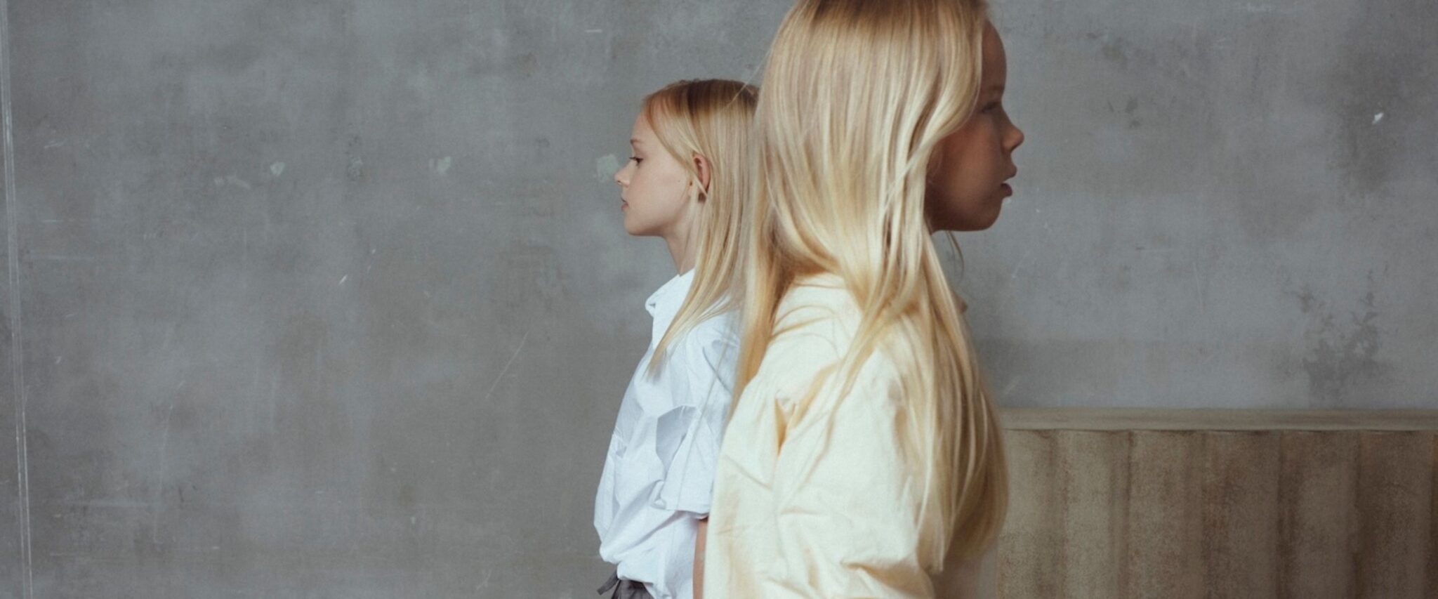 Unlabel Girls SS26 collection – two blonde girls in minimalist pastel outfits standing in profile against a soft grey textured wall, showcasing clean silhouettes and oversized cotton shapes. Scimparello Magazine.