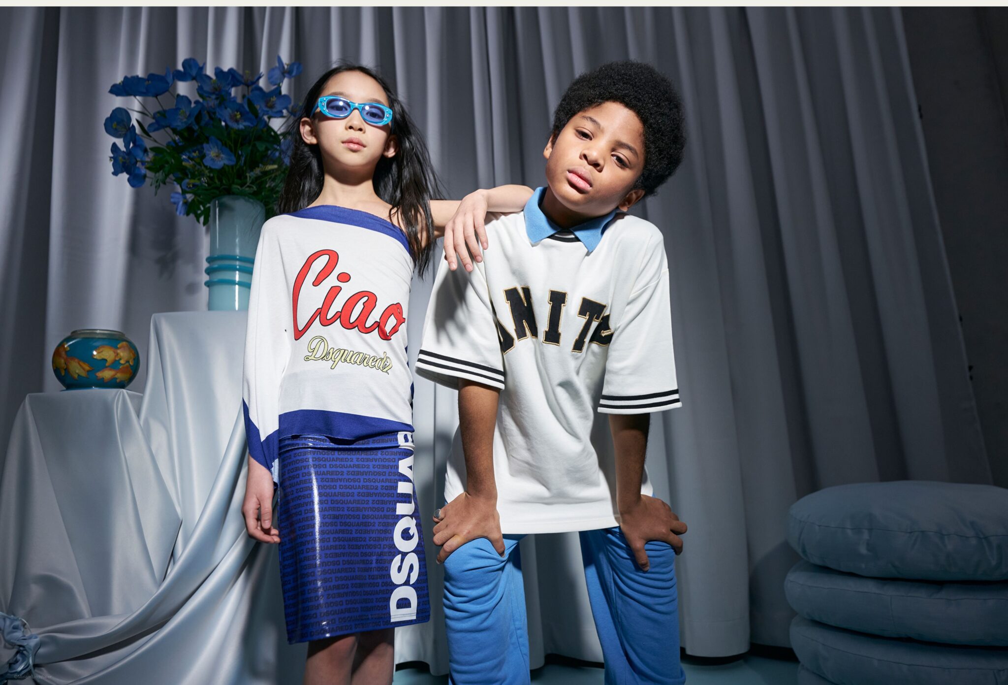 Two children pose together in a studio with soft drapery and blue floral styling; one wears blue sunglasses and a white and blue outfit with bold lettering, while the other leans forward in a sporty white top and blue pants. Photo by Miriam Lindthaler for Scimparello Magazine kids fashion editorial PROM.
