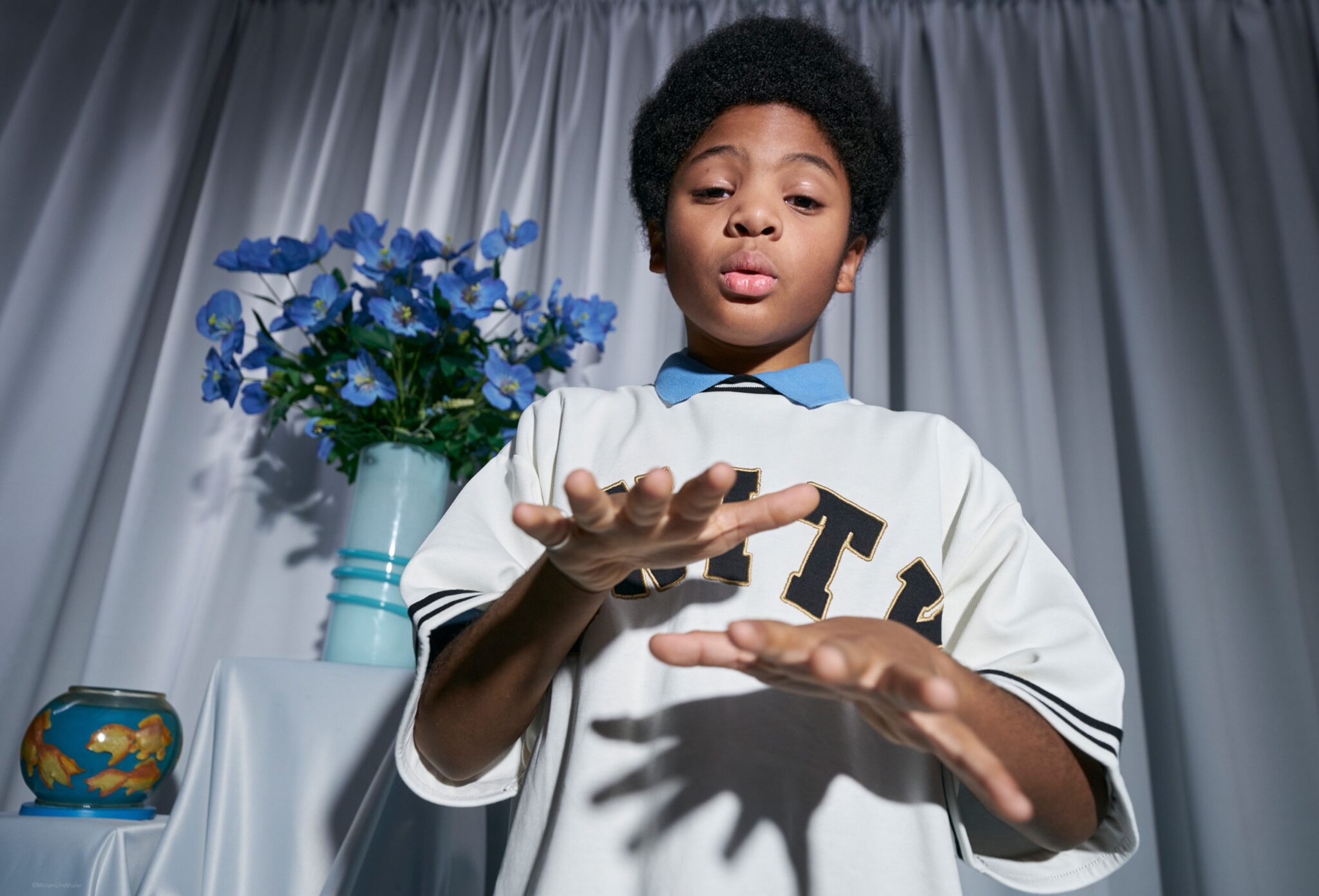 Child in a sporty white shirt with bold lettering gestures with hands in a playful pose, photographed from a low angle in a studio with soft drapery and a vase of blue flowers. Photo by Miriam Lindthaler for Scimparello Magazine kids fashion editorial PROM.
