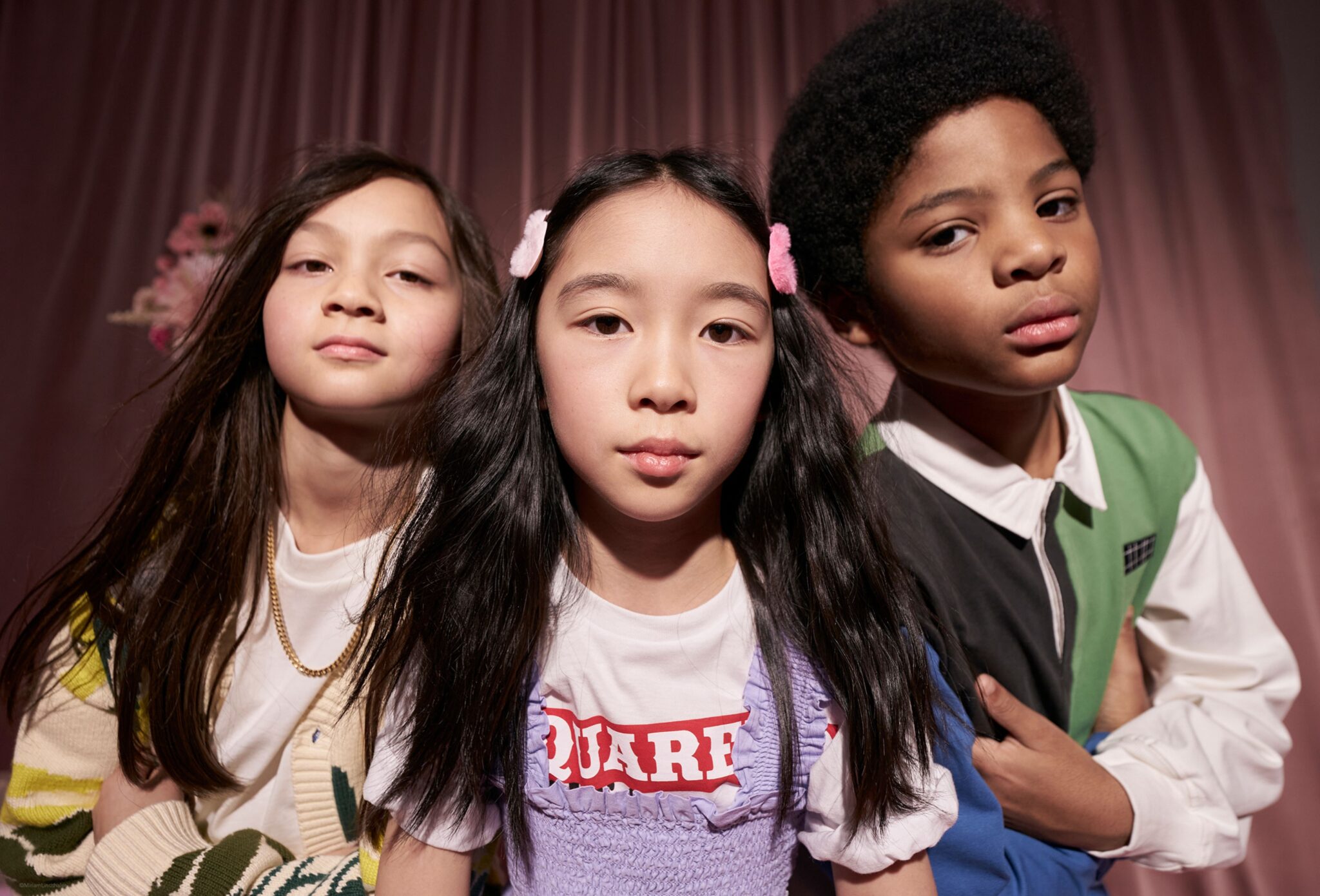 Close-up portrait of three children posing together in a studio with soft pink drapery; styled in layered, colorful outfits with graphic details, they look directly at the camera with confident, expressive faces. Photo by Miriam Lindthaler for Scimparello Magazine kids fashion editorial PROM.