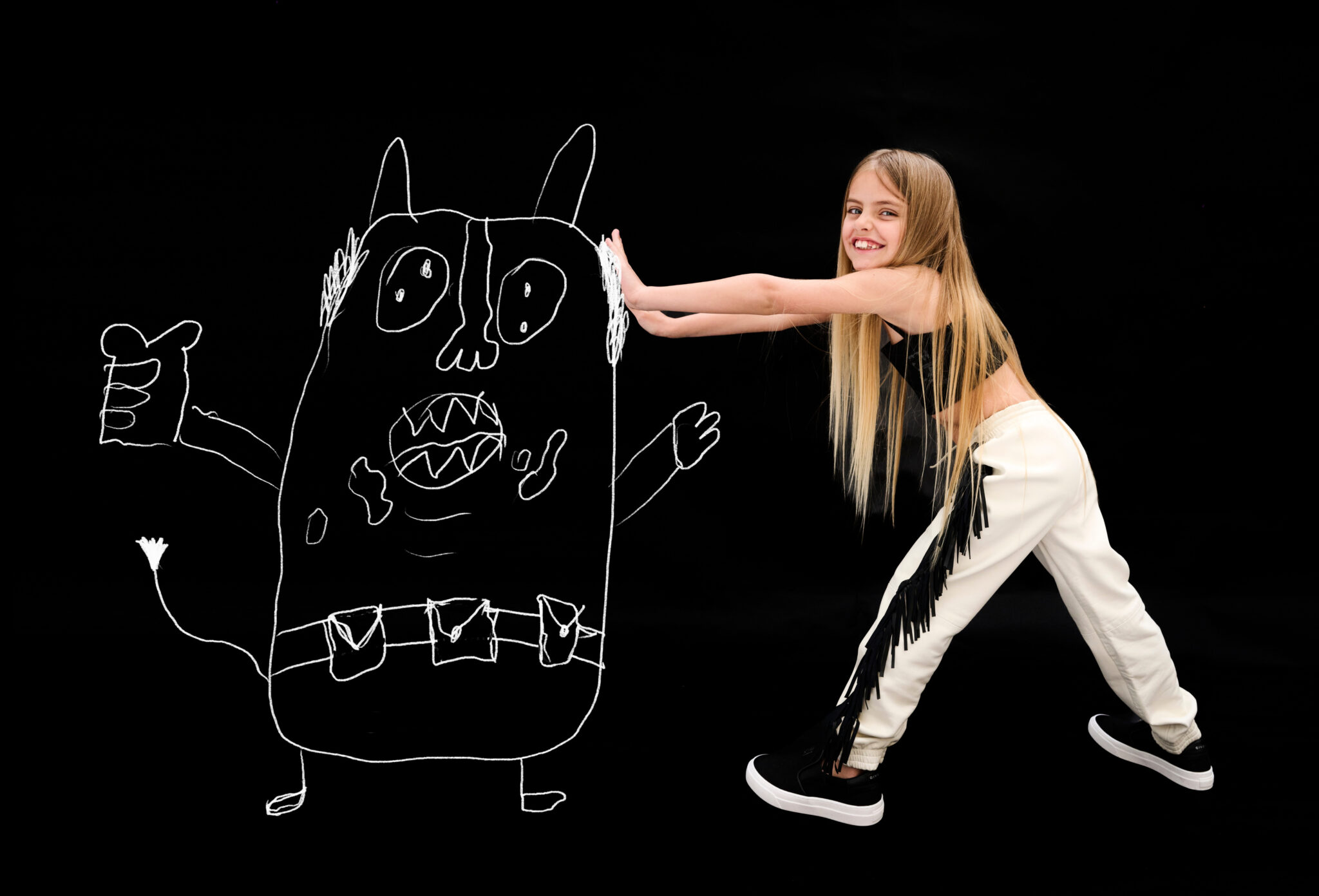 Girl in black top and fringed cream pants pushing against large hand-drawn monster illustration on black background, playful kids fashion editorial DOODLYDOO by Sophie Rata for Scimparello Magazine