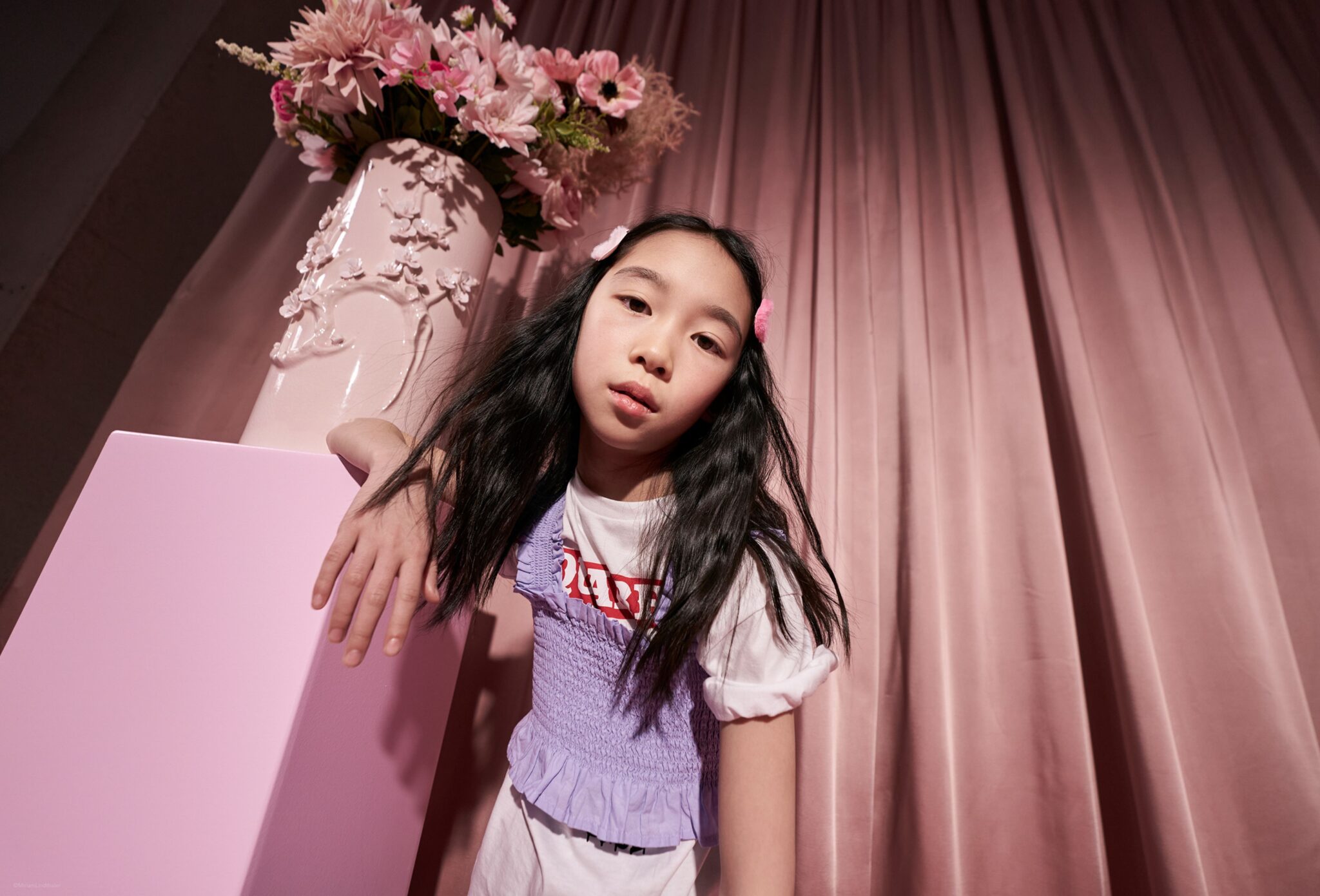 Child leaning against a pastel pink pedestal in a studio setting with draped pink curtains, wearing a white T-shirt layered with a lilac textured top and hair clips, beside a decorative vase filled with pink flowers. Photo by Miriam Lindthaler for Scimparello Magazine kids fashion editorial PROM.