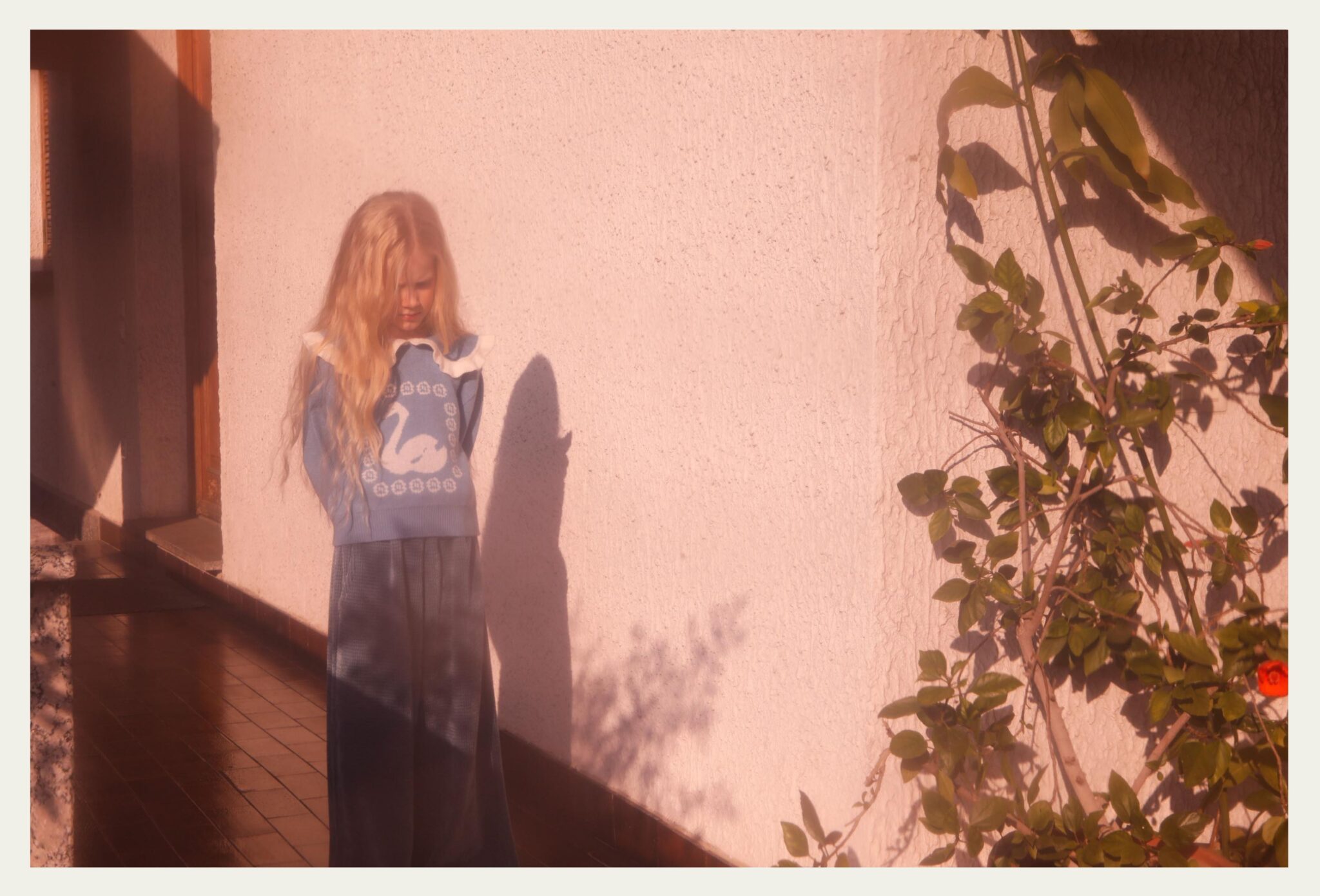 oung girl with long blonde hair stands in warm afternoon light against a textured wall, wearing a blue knit sweater with a swan motif and wide dark trousers, her shadow falling softly beside her near a climbing rose plant. Photo by Annarella Caruso for Scimparello Magazine, kids fashion editorial Sofia, Arthur & Sara.
