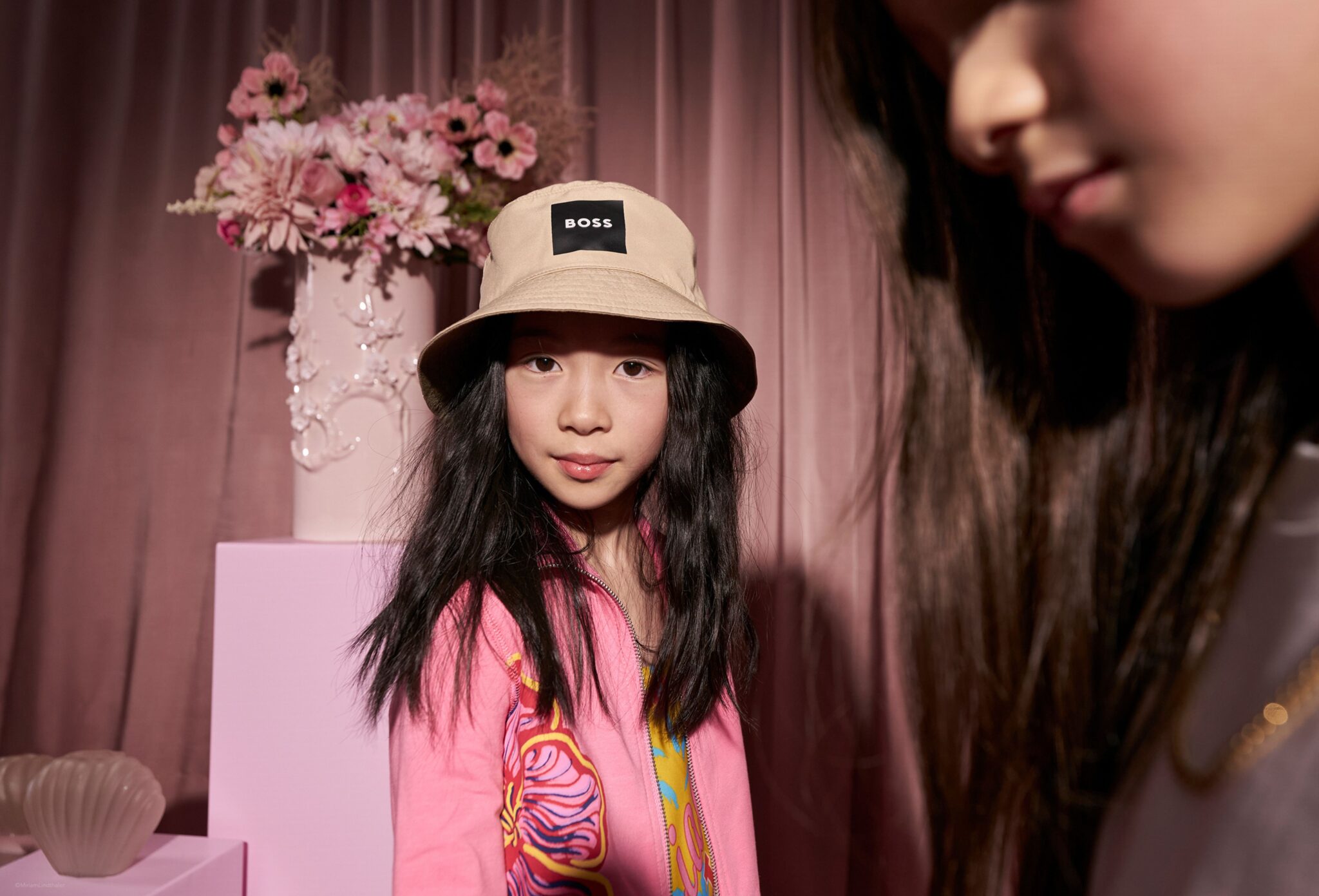 Child wearing a beige bucket hat and a bright pink printed outfit stands in a pink studio setting, looking directly at the camera, with soft drapery and a floral arrangement in the background and another figure blurred in the foreground. Photo by Miriam Lindthaler for Scimparello Magazine kids fashion editorial PROM.