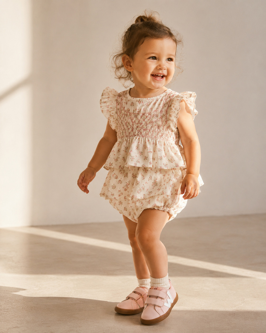 Smiling toddler wearing a romantic floral romper with ruffled sleeves and pink Walkey shoes in soft natural light, Scimparello Magazine.