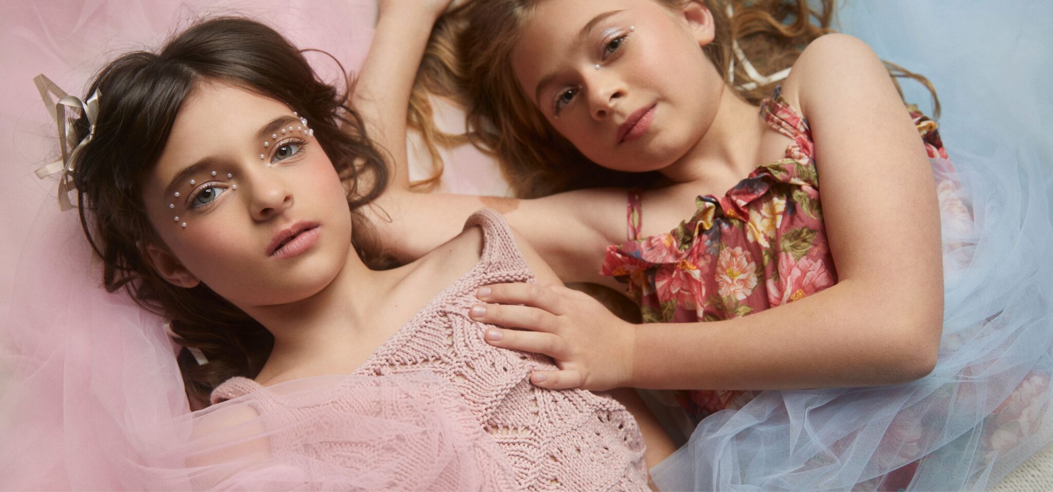 Two girls lying side by side on soft pink and blue tulle, wearing delicate kids fashion looks with floral dress and knitted top, embellished with subtle face gems, dreamy pastel editorial mood, photographed by Chloe Snowdon-Smith, Scimparello Magazine