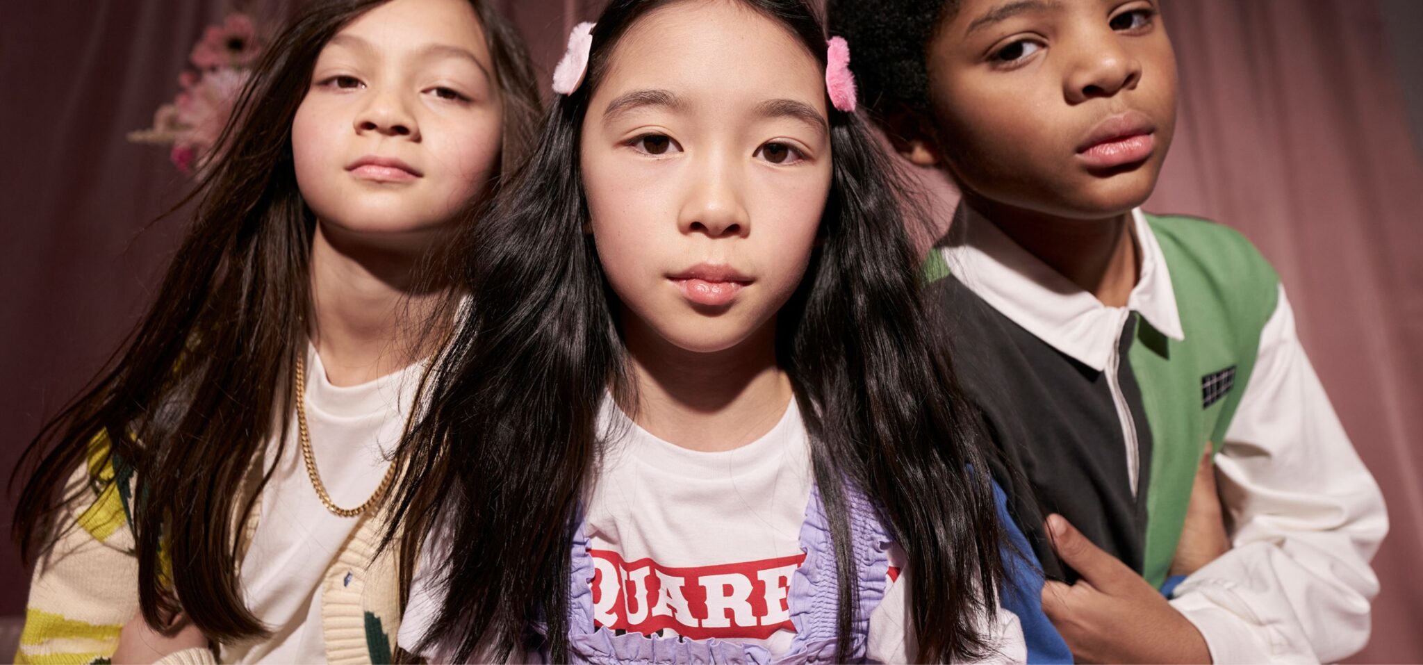 Close-up portrait of three children posing together in a studio with soft pink drapery; styled in layered, colorful outfits with graphic details, they look directly at the camera with confident, expressive faces. Photo by Miriam Lindthaler for Scimparello Magazine kids fashion editorial PROM.
