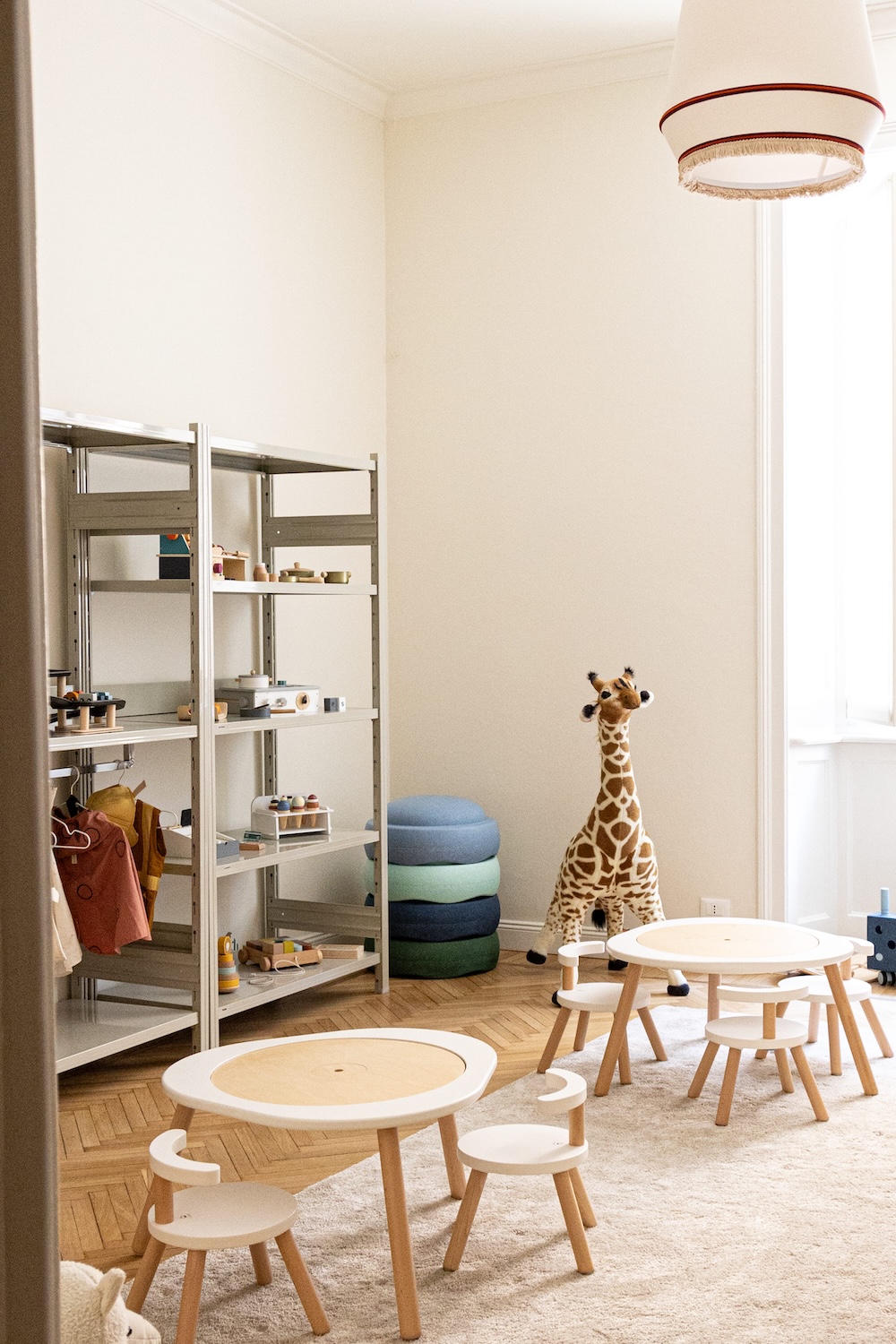 Stylish children’s playroom with round wooden tables and chairs, soft textures, modular shelves and plush giraffe toy in bright Milan apartment setting