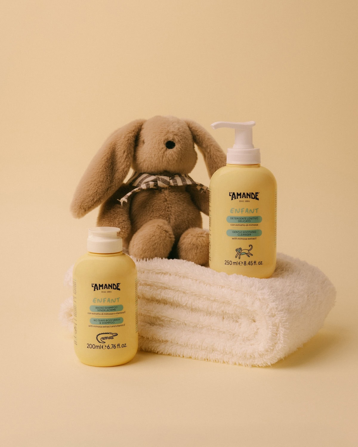 L’Amande Enfant baby skincare products including gentle cleanser and no-tears shampoo, displayed with a soft towel and plush bunny on a pastel background.