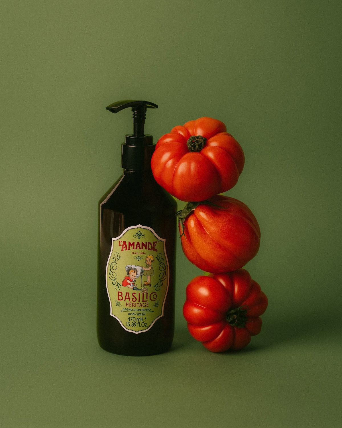 L’Amande Basilico Heritage body wash bottle with fresh tomatoes, highlighting natural Italian ingredients on a green background.