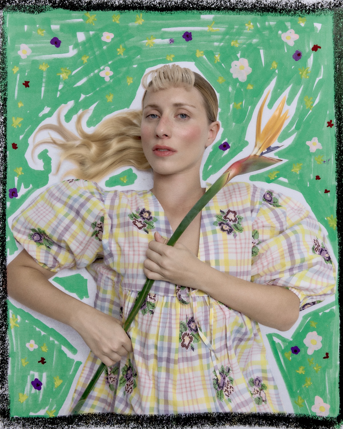 Blonde woman lying on a hand-painted green floral background, wearing a pastel plaid dress with embroidered flowers and holding a tropical bird-of-paradise stem, evoking playful, childlike summer fashion.