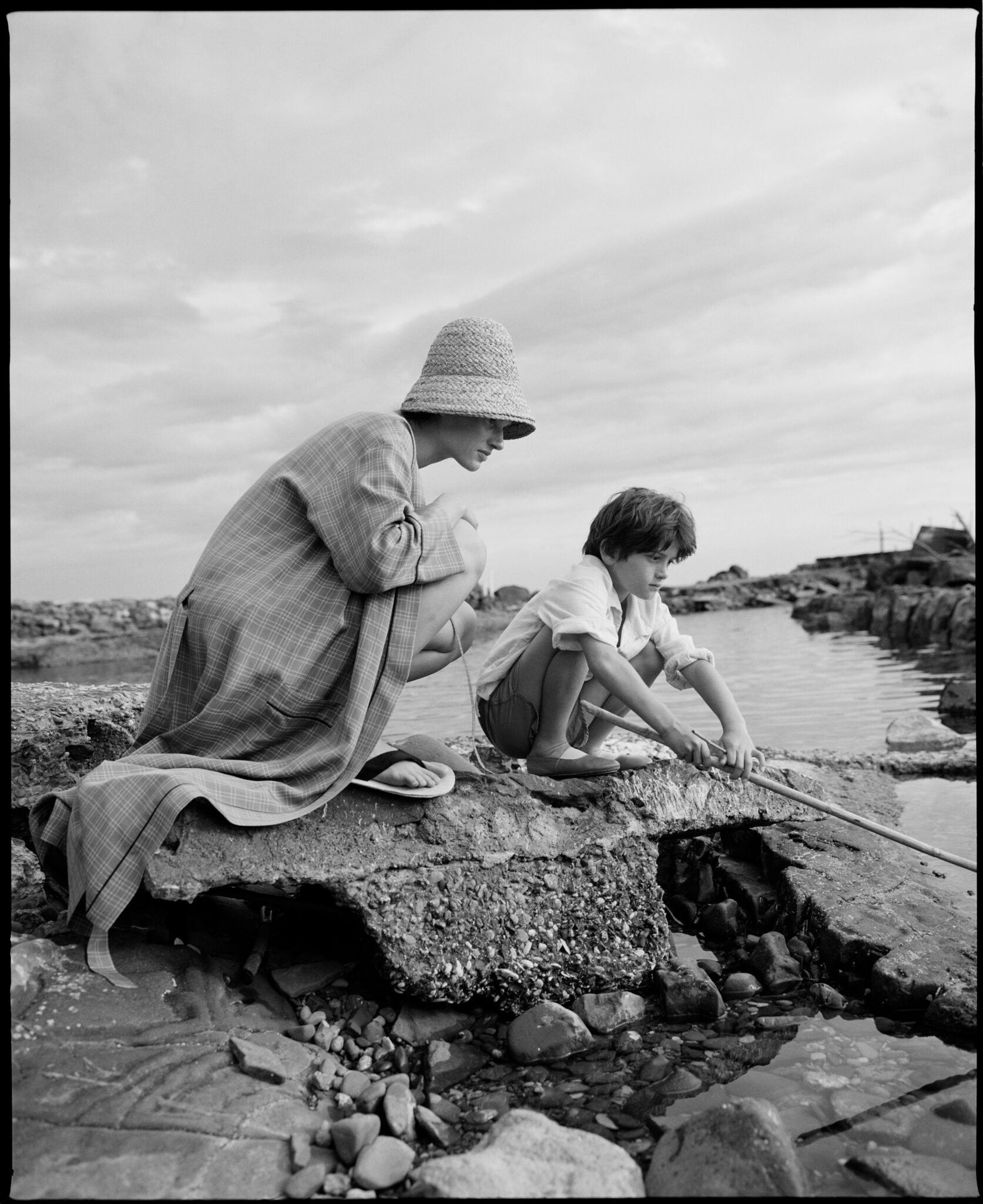 Black and white campaign image showing a woman in a checked coat and woven hat sitting beside a young boy by the rocky seaside, wearing pieces from the Caramel x Zara collection.