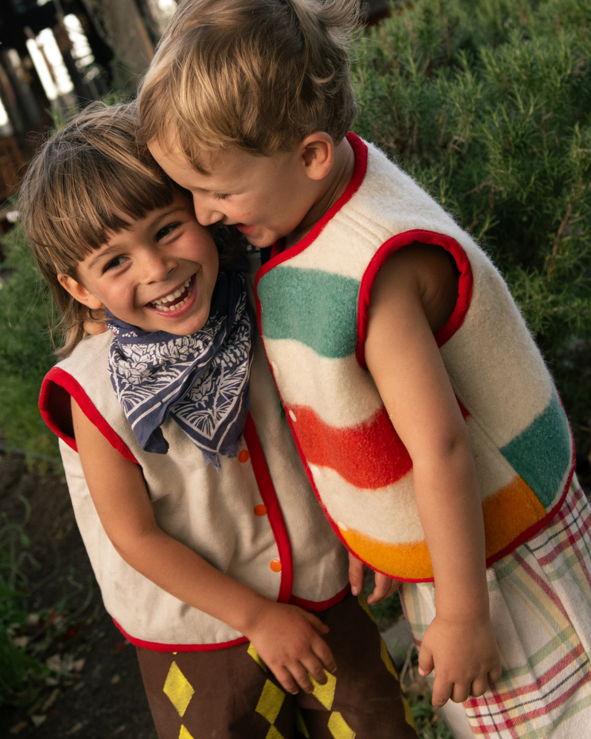 Two smiling children wearing handcrafted Playdate Vintage wool vests with colorful striped details and playful patterns, photographed outdoors for Scimparello Magazine.