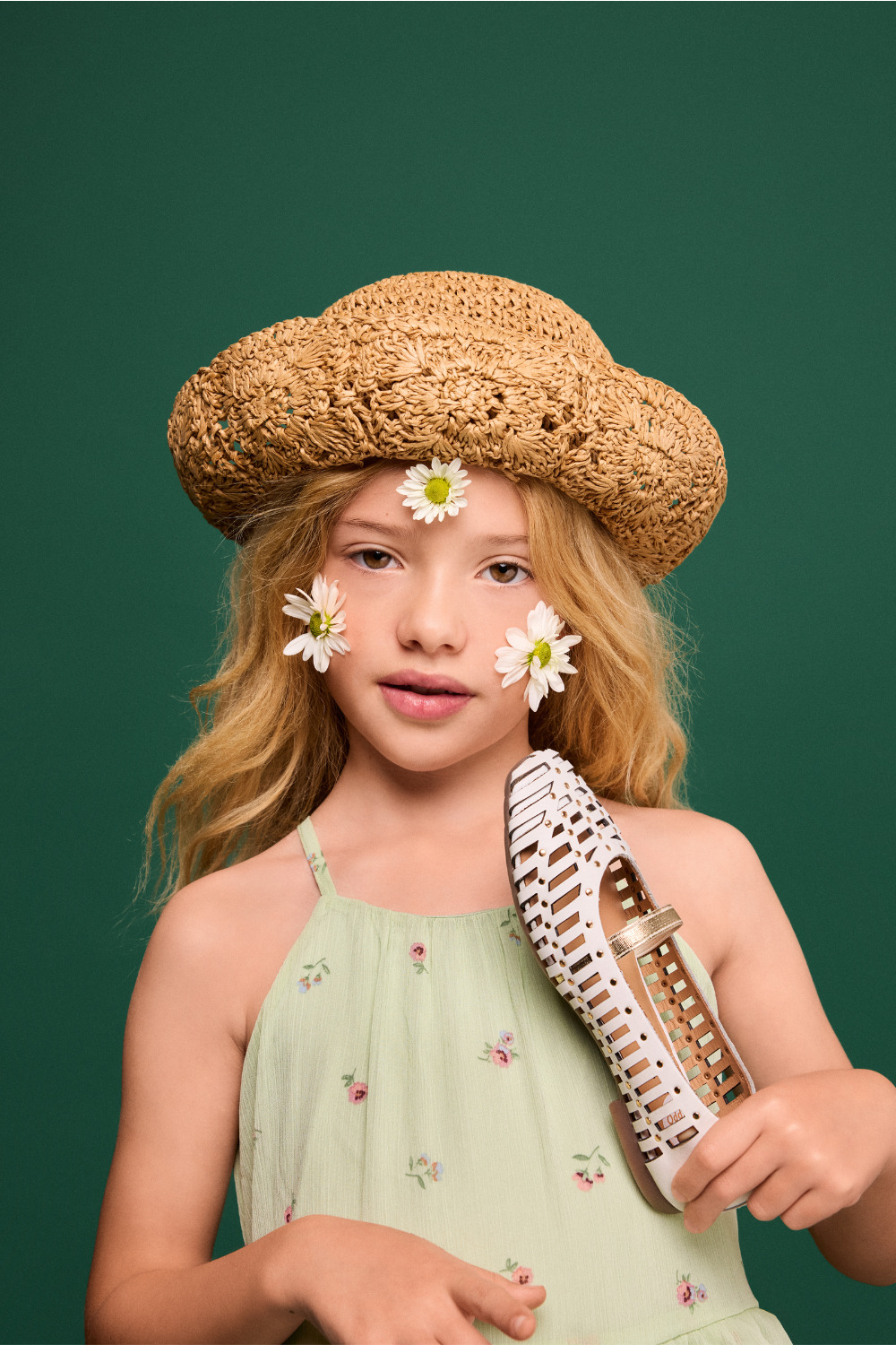 Girl wearing a straw hat and pale green floral dress, holding a white Gioseppo Kids SS26 shoe with daisy flowers on her face for Scimparello Magazine.