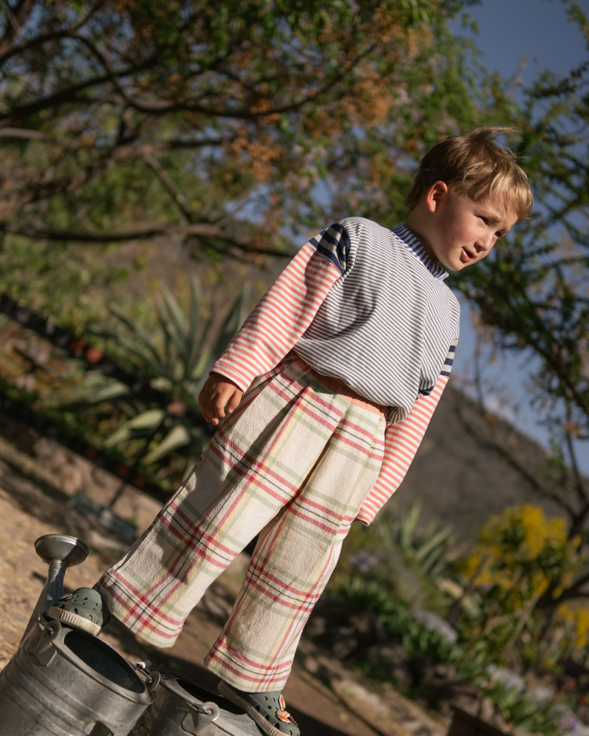 Child wearing striped top and plaid trousers from Playdate Vintage standing outdoors in a sunny garden setting for Scimparello Magazine.
