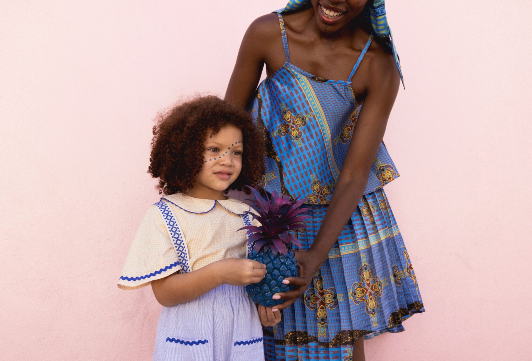 Adult and child models in coordinated blue patterned outfits standing against a soft pink wall, holding a painted pineapple, playful mix of color and cultural details, kids fashion editorial Culture Clash by Hannah Mentz shot in South Africa for Scimparello Magazine