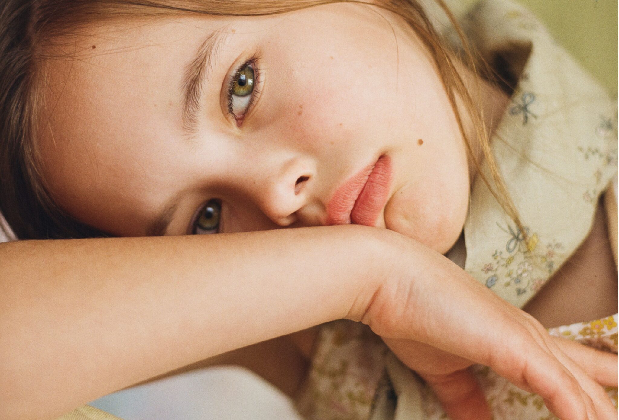 Close-up portrait of a young girl resting her face on her arm, wearing a delicate floral kids fashion look in soft vintage tones for Scimparello Magazine editorial Printed Heirlooms by Emma Wright.
