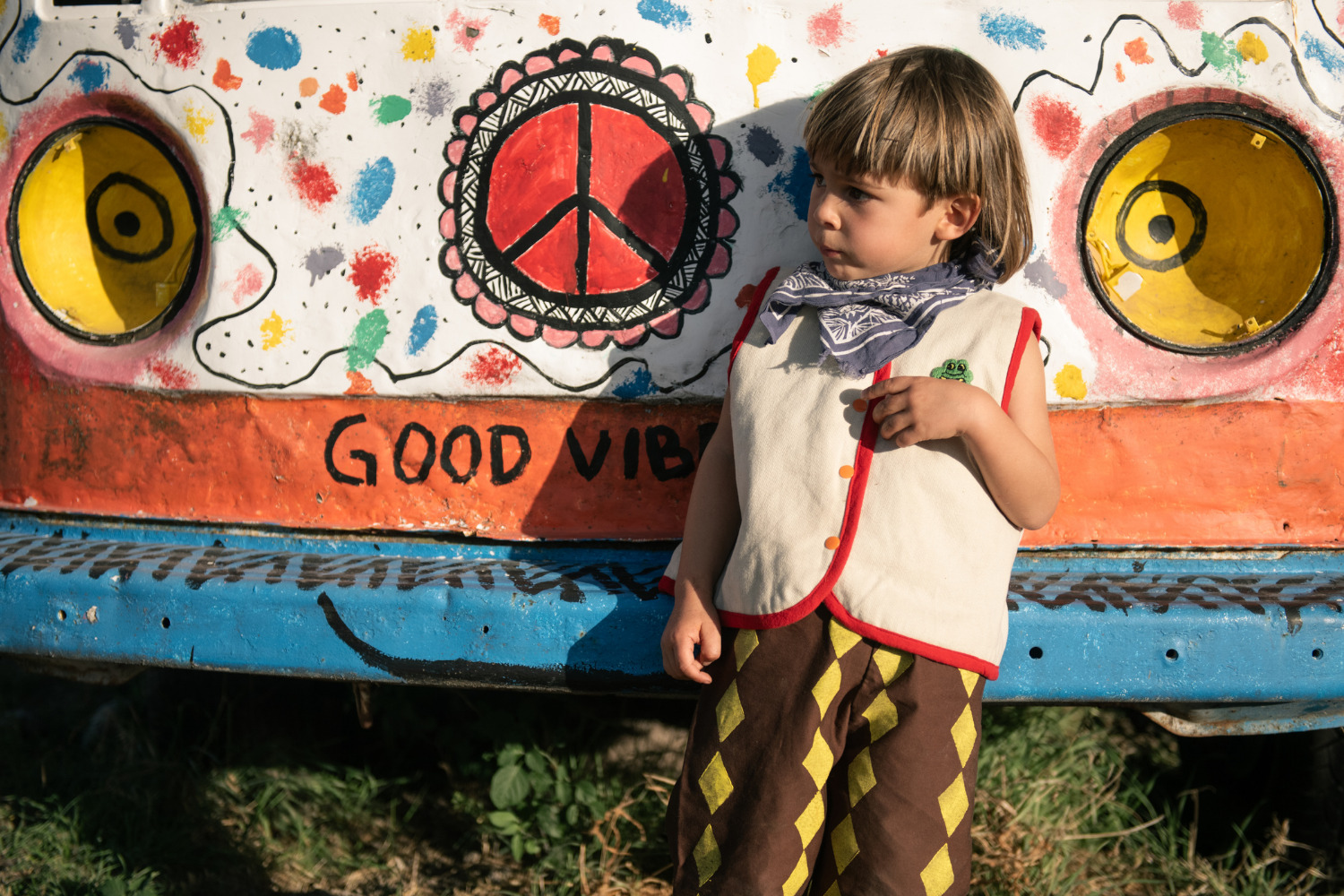 Child wearing a handmade Playdate Vintage vest and patterned trousers standing in front of a colorful painted wall with peace sign artwork and “Good Vibes” text for Scimparello Magazine.