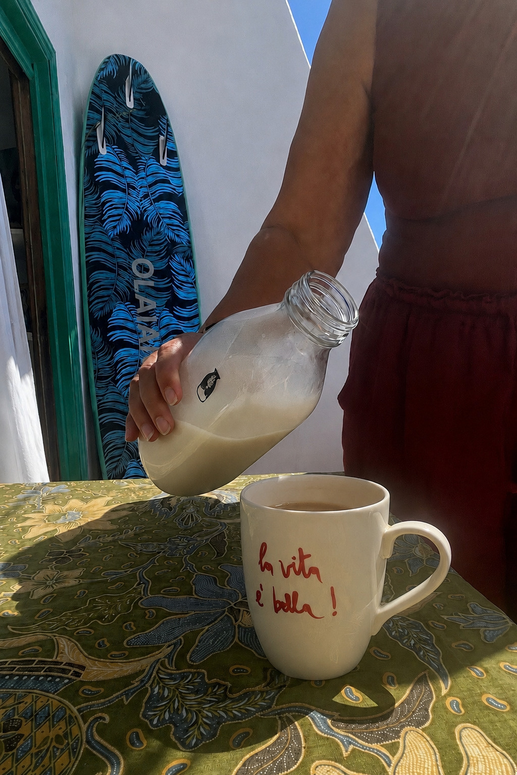 Homemade plant milk being poured from a glass bottle into a white mug with the phrase “la vita è bella” on a patterned table outdoors in Lanzarote. A blue surfboard leans against the wall in the background. Scimparello Magazine lifestyle image.