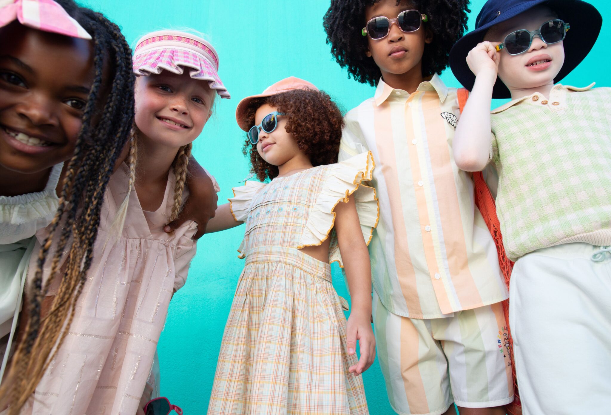 Group of children posing together in pastel-toned outfits and sunglasses against a turquoise wall, playful attitude and summer styling with soft colors and textures, kids fashion editorial Culture Clash by Hannah Mentz shot in South Africa for Scimparello Magazine
