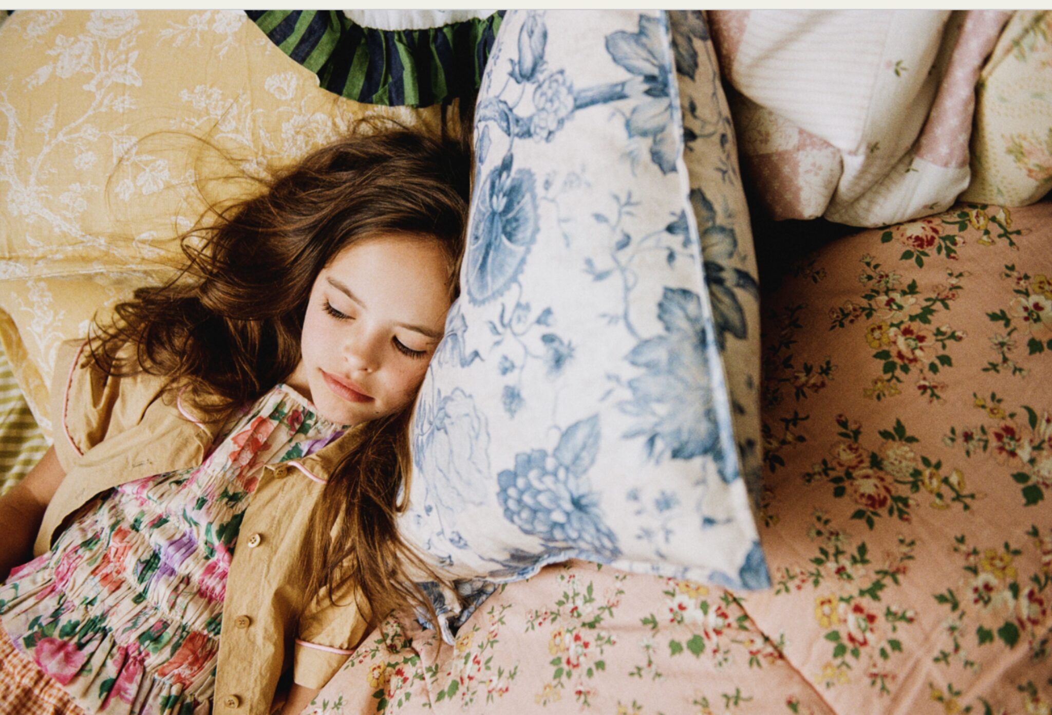 Girl rests among layered floral pillows and vintage patterned textiles, wearing a floral kids fashion look with a soft tan jacket for Scimparello Magazine editorial Printed Heirlooms by Emma Wright.