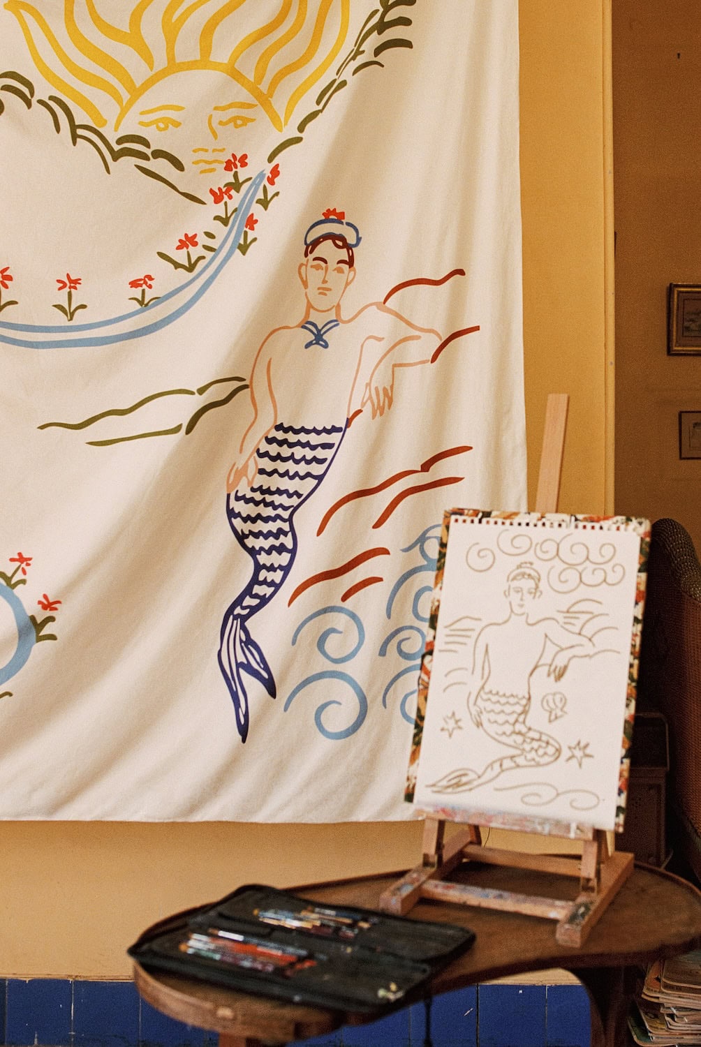 Mythological mermaid-inspired illustration from the Bonsoirs x Jacques Merle collaboration displayed on a large textile backdrop, alongside an artist sketch on an easel in a warm Mediterranean-style interior.