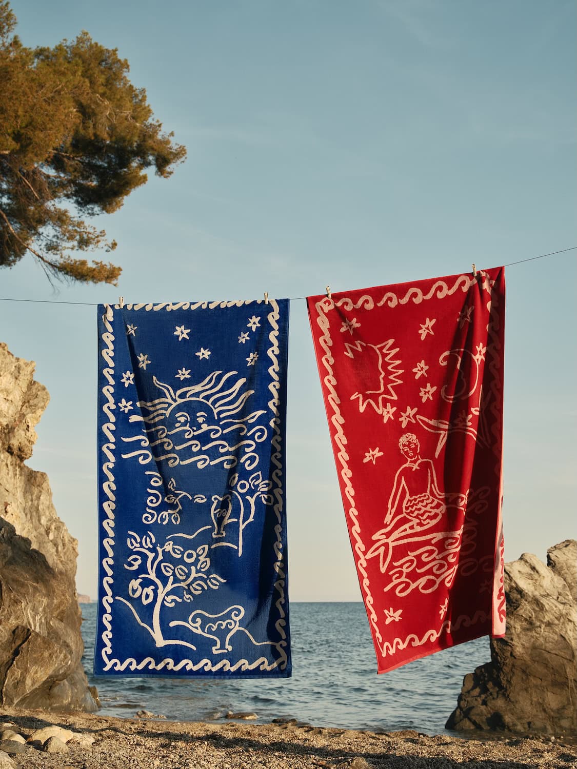 Two illustrated beach towels from the Bonsoirs x Jacques Merle collaboration hanging on a clothesline by the sea, featuring whimsical Mediterranean-inspired drawings in deep blue and vibrant red tones against a sunny coastal landscape.
