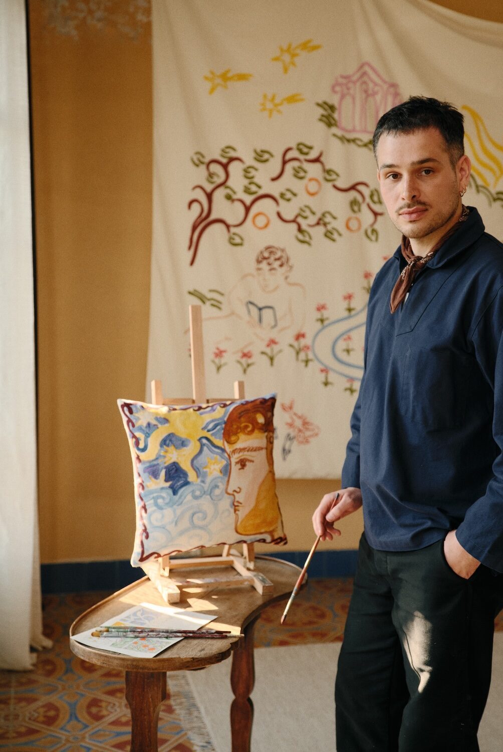 French painter Jacques Merle standing beside a hand-painted cushion from the Bonsoirs collaboration, surrounded by whimsical Mediterranean-inspired illustrations, embroidered textiles and warm sunlit interiors.