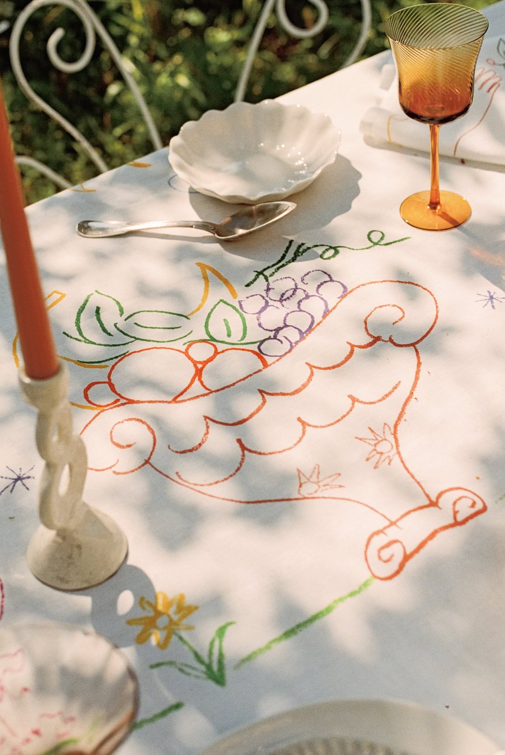 Artistic table setting from the Bonsoirs x Jacques Merle collaboration featuring whimsical hand-drawn motifs, embroidered-style illustrations and warm summer light in a garden setting.