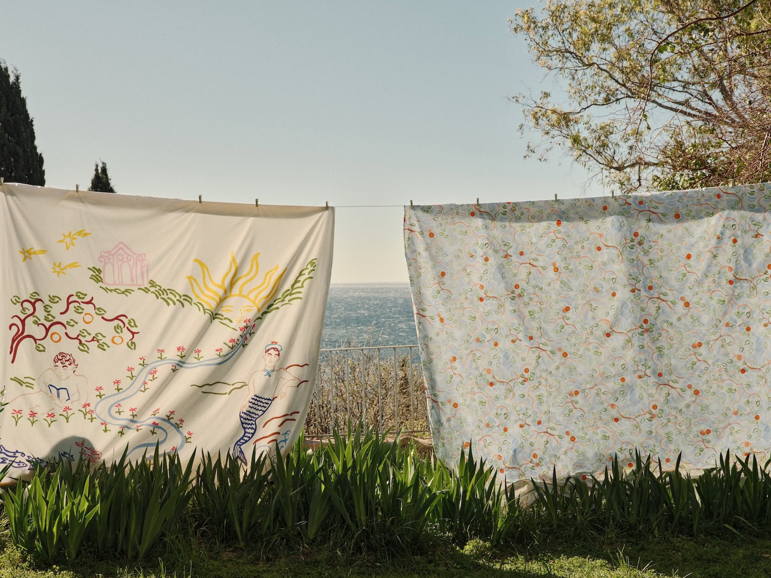 Illustrated Bonsoirs x Jacques Merle textiles hanging outdoors by the sea, featuring whimsical Mediterranean-inspired drawings, mythological figures and sun motifs in a dreamy French Riviera setting.