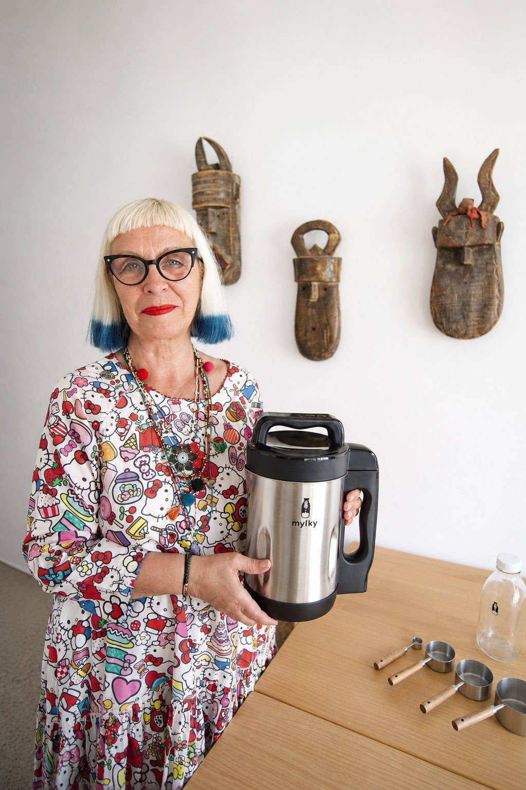 Petra Barkhof holding the Mylky plant milk maker at home, wearing a colorful Hello Kitty print top and signature blond hairstyle with blue tips. Lifestyle portrait for Scimparello Magazine.