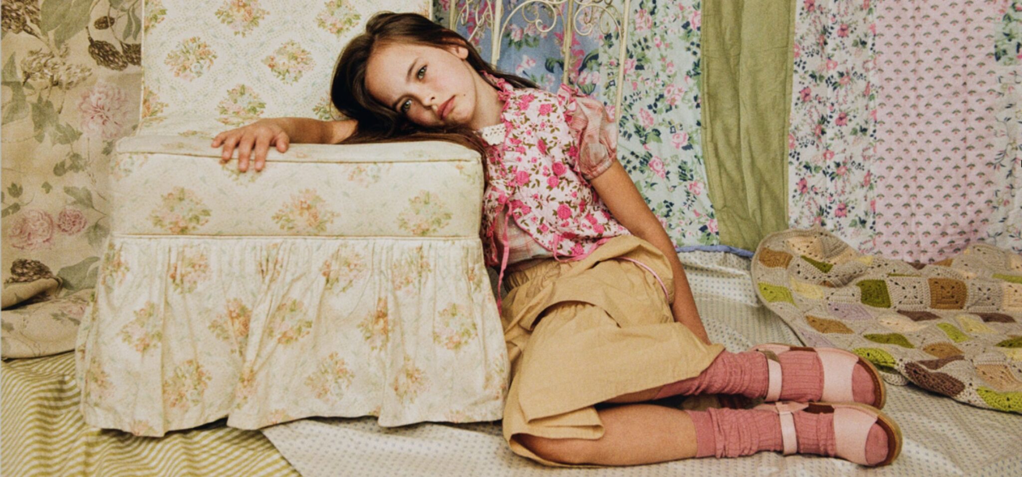 Young girl in a floral kids fashion look sits beside a vintage floral armchair in a room filled with layered printed fabrics, crochet details and soft nostalgic colors for Scimparello Magazine editorial Printed Heirlooms by Emma Wright.