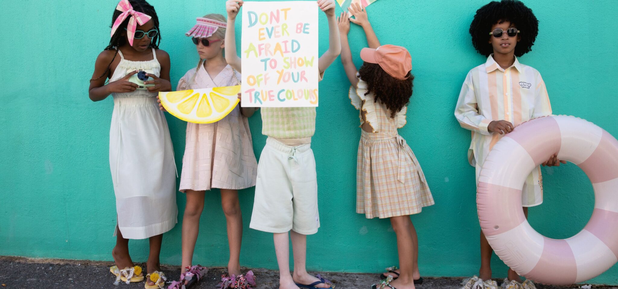 Group of children posing together in pastel-toned outfits and sunglasses against a turquoise wall, playful attitude and summer styling with soft colors and textures, kids fashion editorial Culture Clash by Hannah Mentz shot in South Africa for Scimparello Magazine