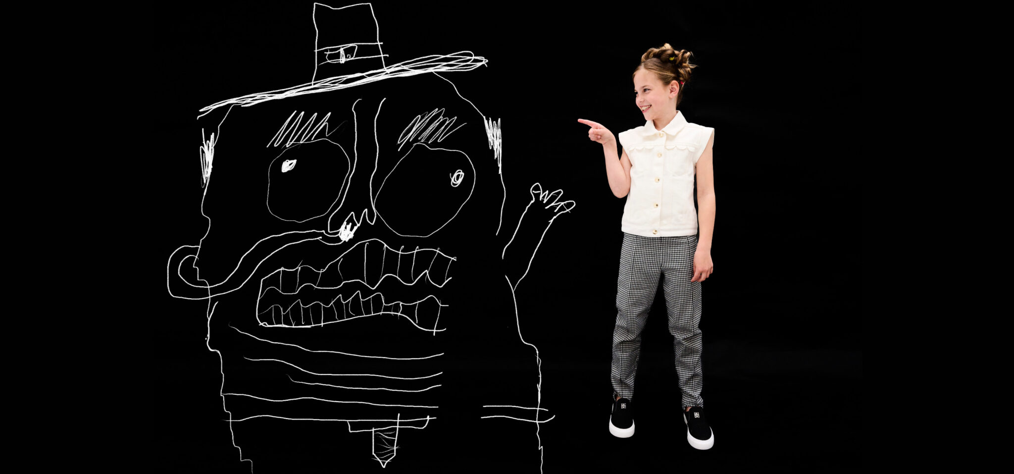 Girl in white blouse and checkered pants pointing at large hand-drawn monster with hat on black background, playful kids fashion editorial DOODLYDOO by Sophie Rata for Scimparello Magazine
