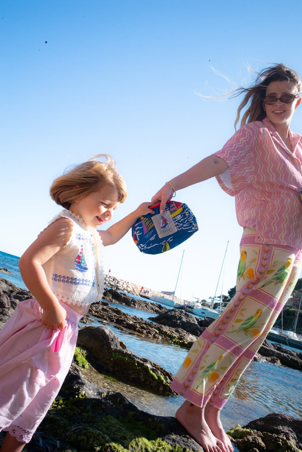 Child and woman walking across seaside rocks on a sunny day, holding a quilted blue pouch from the Maison Mangostan x Studio Noos capsule collection. The scene captures a carefree Mediterranean summer atmosphere with boats and crystal-clear water in the background.