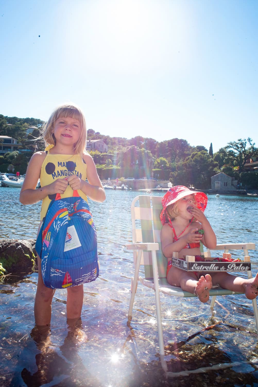 Two children enjoying a sunny day by the sea, surrounded by shallow water and carrying a quilted blue backpack from the Maison Mangostan x Studio Noos collection. The colorful accessories feature playful sailboat prints inspired by summer adventures.