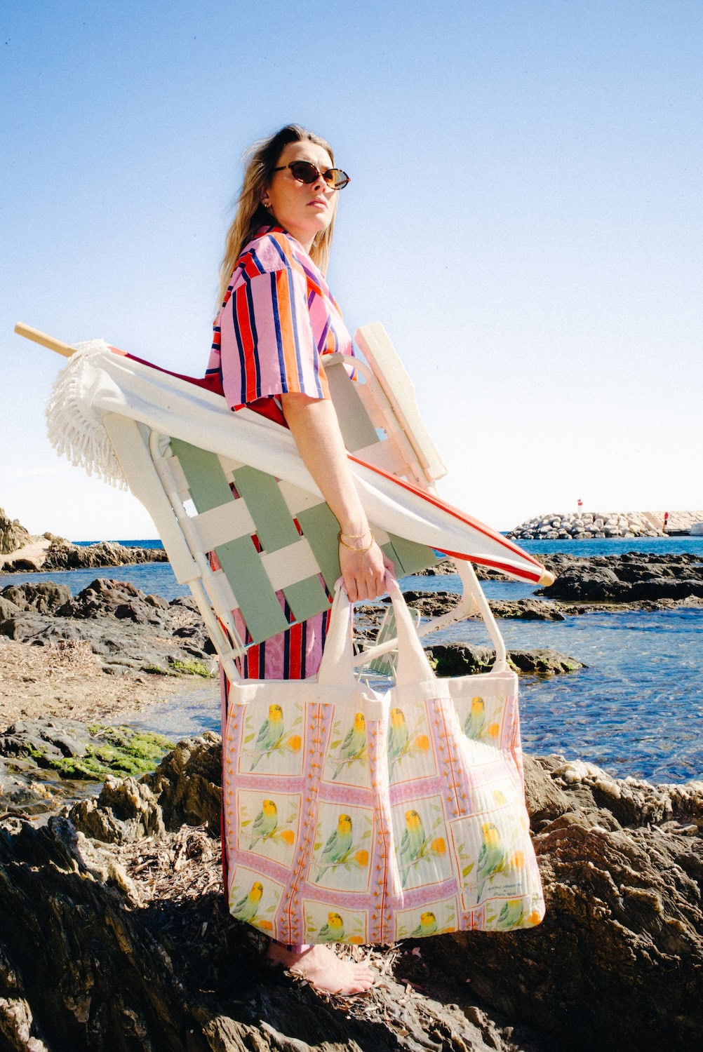 Woman carrying a beach chair and a large quilted tote bag featuring tropical bird prints from the Maison Mangostan x Studio Noos capsule collection. Set against a rocky Mediterranean coastline, the image evokes effortless summer escapes.
