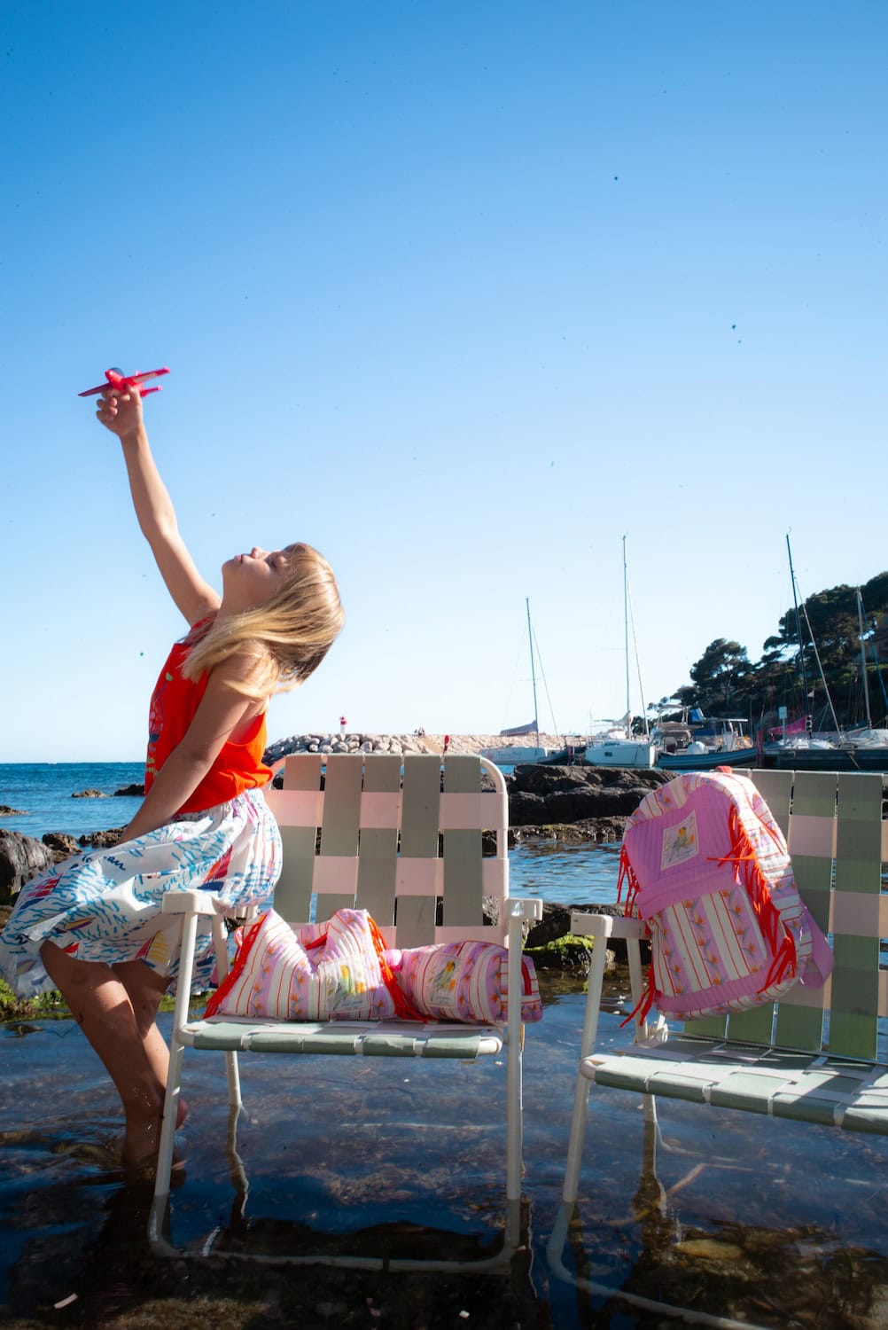 Child playing by the water with a toy airplane beside quilted bags and a backpack from the Maison Mangostan x Studio Noos capsule collection. Bright colors, seaside scenery and whimsical prints celebrate the joy of summer exploration.